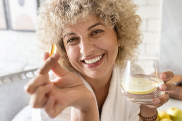 Woman Healthcare. Female Holding Suplement in a Bright Room. Portrait of a joyful curly-haired woman taking a vitamin pill with a glass of water. Concept of health, wellness, and daily routine  - 741314605