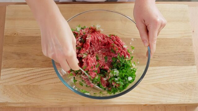 The chef mixes minced meat, beef, pork, chicken, turkey with the addition of onions, spices and herbs in a glass bowl with a spoon, wooden table. Close-up, top view