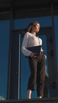 An attractive dark-haired businesswoman in a white shirt with a laptop in her hands is walking near a glass business center.Vertical video.