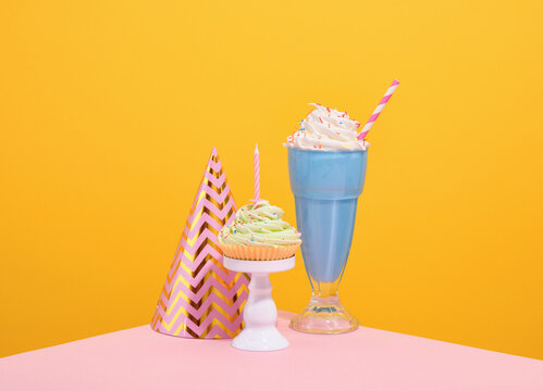 Sweet desserts and a holiday hat. Blue milkshake with bright sprinkles and straw, cupcake with whipped cream and birthday candle.