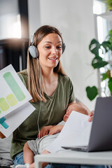 Young new mom holding cute baby in her arms at home while working on a computer