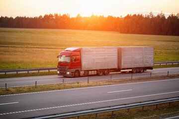 A truck with a semi-trailer and a hitch transports cargo against the backdrop of a sunset along the...
