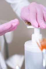 Cosmetic Gel Dispensing for Beauty Treatments. Gloved professional cosmetologist's hands dispensing a clear gel from a white pump bottle, with a soft focus on cosmetic brushes in the background. 