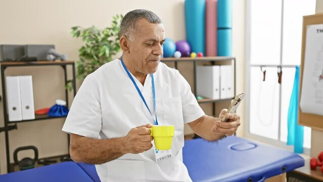 Smiling middle-aged hispanic man in a white coat holding a mug and smartphone in rehab clinic interior.