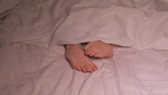 kid boy legs itching and scabies in bed in the bedroom in the evening. Restless baby child girl scratches her feet with fungus on the sheet under the blanket