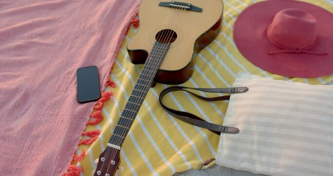 A guitar lies next to a hat and phone on a beach blanket