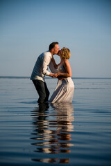 beautiful  happy couple, bride and groom in white dresses walking in the ocean on sunny summer day and holding each other hand by hands