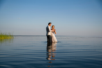 beautiful  happy couple, bride and groom in white dresses walking in the ocean on sunny summer day and holding each other hand by hands