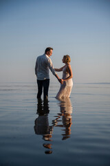 beautiful  happy couple, bride and groom in white dresses walking in the ocean on sunny summer day and holding each other hand by hands