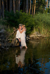 beautiful hugging couple, bride and groom in white dresses sitting on the  stump  in forest lake on sunny summer day