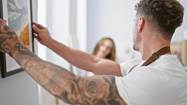 A tattooed man and spectacled woman collaborate in hanging a framed painting in a bright art studio.