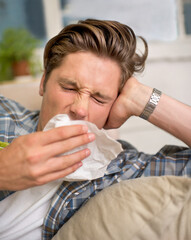 Sick, flu or man sneezing into tissue on sofa with cold, virus or hay fever at home. Influenza,...