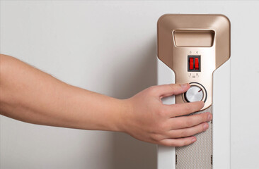 A man's hand regulates the heating power of an electric air heater in an apartment, close-up. Copy space for text, radiator
