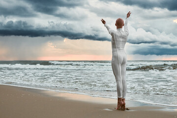 Hairless girl with alopecia in white futuristic suit standing on sea beach stretched out arms to cloudy sky, full length back view portrait, metaphoric performance with bald sensitive teenage girl
