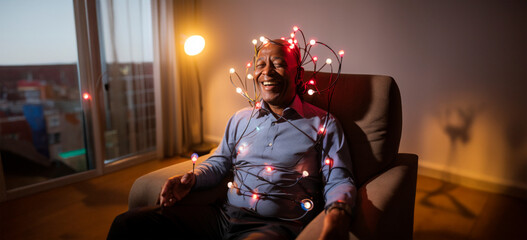 Joyful elderly man wrapped in colorful Christmas lights, sitting in a cozy room, embodying festive spirit and happiness.