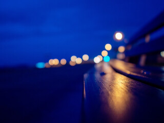 City lights illuminate wooden bench. Blue night sky with town lights out of focus in the...