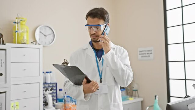 A Focused Hispanic Man In Lab Coat Consulting Information On Clipboard While Talking On The Phone In A Lab