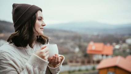 Young woman in white knitted sweater holding white cup of tea, standing on a balcony of a wooden house with mountains view.