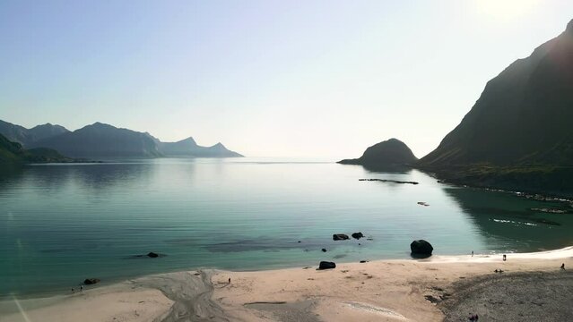 From the sandy shores of Haukland Beach, a drone ascends, showcasing the stunning contrast of white sands and emerald waters within a secluded bay in Lofoten, Norway