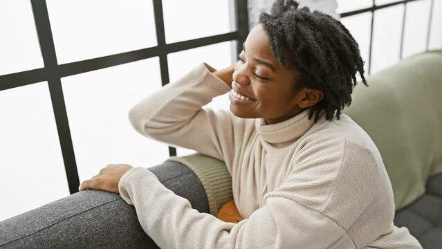 Confident young black woman with fashionable dreadlocks joyfully posing on the cozy sofa at home, touching hair and smiling cheerfully.