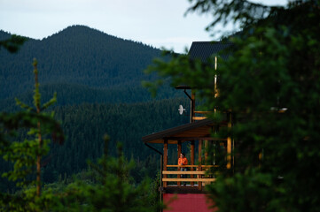 a girl in an orange jacket stands on the terrace of a wooden house against the background of...