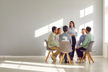 Corporate business team meeting with a manager or coach. Group of happy diverse, multiracial people sitting in a circle around a standing woman in an office with a light grey copy space wall - obrazy, fototapety, plakaty