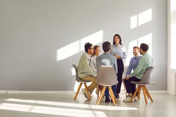 Fotobehang Corporate business team meeting with a manager or coach. Group of happy diverse, multiracial people sitting in a circle around a standing woman in an office with a light grey copy space wall © Studio Romantic