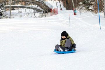 A boy rides a sleigh. A boy in a winter jacket goes down a hill.