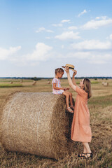 Mom and little daughter in a field of harvested wheat against the blue sky. The daughter is sitting on a bale of straw. Mom puts a straw hat on her daughter