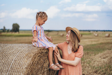 Naklejka na ściany i meble A young mother in a straw hat and a young daughter communicating on a harvested wheat field, a girl sitting on a bale of straw