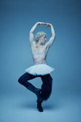 Shirtless man in tutu and boots posing in ballet stance against grey studio background, embodying...