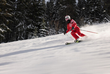 Female skier in a vibrant red ski suit skiing a snowy slope. Trees lining the run are frosted with snow. Cold environment typical of a winter sports setting. 
