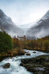 Norwegian mountain landscape, cloudy, with an autumn forest on a river, with the tongue of the Briksdalsbreen glacier in the background, over the snow-capped mountains.