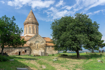 Church of Khirsa monastery of Saint Stephen. Stone walls, orange tile roof, green lawn, blue bright sky with clouds, gravestones and huge tree. Visible Alazani valley
