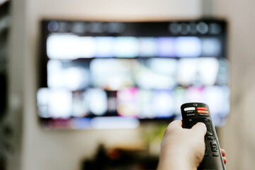 Hand holding remote control pointing to TV. Person watching smart television indoors. Blurred background. Colorful screen. Streaming series, channels. Woman browsing, looking for films online 