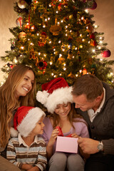 Fototapeta na wymiar Christmas, tree and family with gift, celebration together and lights for happy surprise at night. Father, mother and children smile in living room for present giving, festive xmas or love in home.