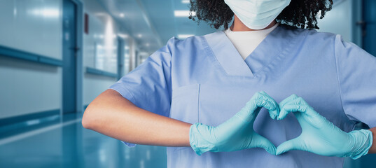 A healthcare professional in a blue scrub top and protective mask makes a heart shape