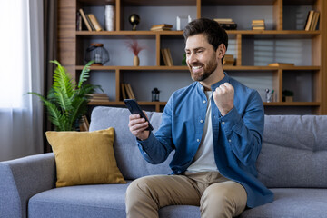 Smiling man enjoying success while using smartphone on couch at home