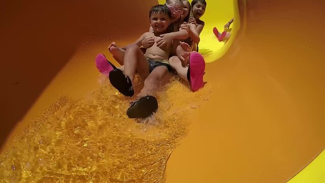 Laughing children ride together water slide on sunny day. Slow motion