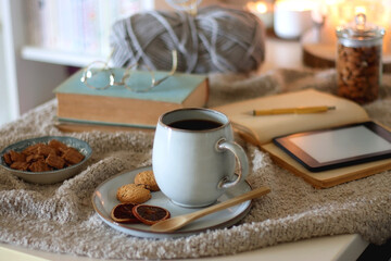 Cup of tea or coffee, cookies, books, glasses, e-reader, almonds, ball of yarn, knitting needles and lit candles on the table. Hygge at home. Selective focus.