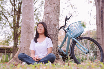 Fototapeta na wymiar 12 year old Asian girl wearing eyeglasses and a white shirt sitting meditating under a tree in the park with blue bicycle, Young woman with Mental health and relaxation in nature concept.