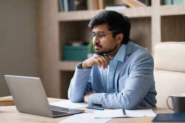 Thoughtful Indian businessman pondering strategies reviewing work on laptop indoors