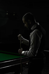 Dark image of young man in classical wear preparing cue with chalk before snooker game. Promotional...