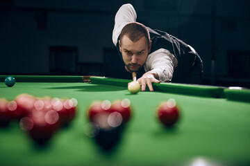 Concentrated young man leaning on billiards table, playing snooker game. Lifestyle magazine spread highlighting leisure activities. Concept of billiards sport, gambling, hobby, leisure, game - obrazy, fototapety, plakaty