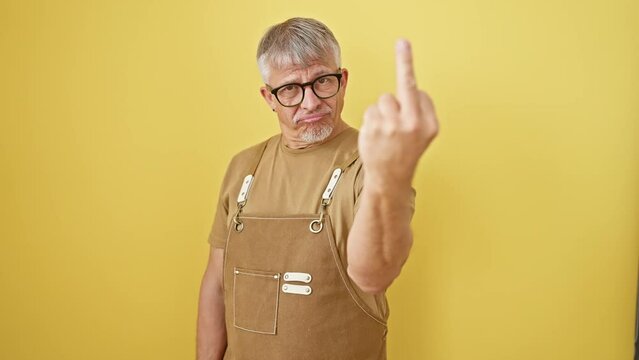 Impudent middle-age grey-haired man in glasses and apron standing, showcasing a rude 'fuck you' hand gesture. angry expression over isolated yellow wall background