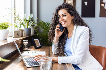 Smiling pretty skilled young woman with wireless earbuds sitting at table in office and working with modern computer. Happy businesswoman taking a video call in a co-working space