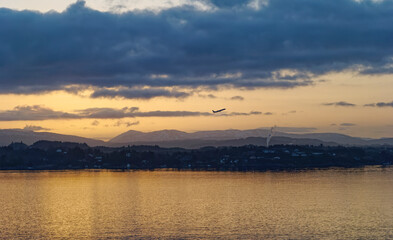 An Aircraft taking off from Bergen Airport at dawn passing over isolated Houses at the water edge of the Fjord under low cloud.