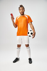 shocked african american man holding phone and football and looking at camera, online betting