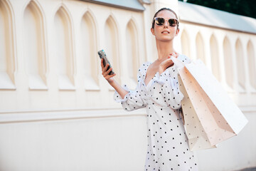 Young beautiful smiling hipster woman in trendy summer clothes. Sexy carefree model walking in the street background at sunset. Positive female holding shopping bag. Posing outdoors. Holds smartphone