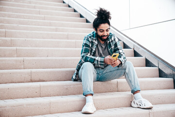 Handsome smiling hipster model. Unshaven Arabian man dressed in summer casual clothes. Fashion male with long curly hairstyle posing in the street. Holds smartphone, uses phone mobile apps - obrazy, fototapety, plakaty